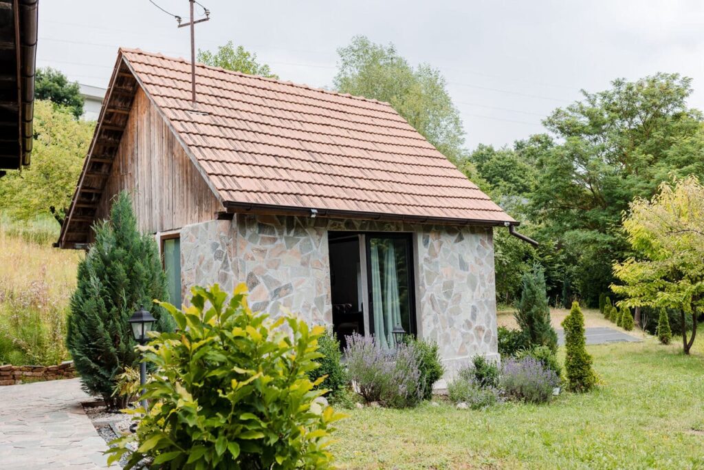 O vedere laterală a noii anexe a Cabanei Ana, cu pereți din piatră rustică și lemn, acoperiș de țiglă și o terasă cu ușă de sticlă. Clădirea este înconjurată de verdeață luxuriantă, arbuști și alei de piatră.