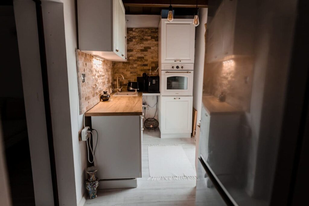 Vedere din living spre bucătăria Cabanei Ana. Se observă o bucătărie modernă, cu mobilier alb, o masă de lucru din lemn, un perete decorativ cu piatră, cuptor încorporat și un frigider. Bucătăria este curată și bine luminată, sugerând că este complet utilată.