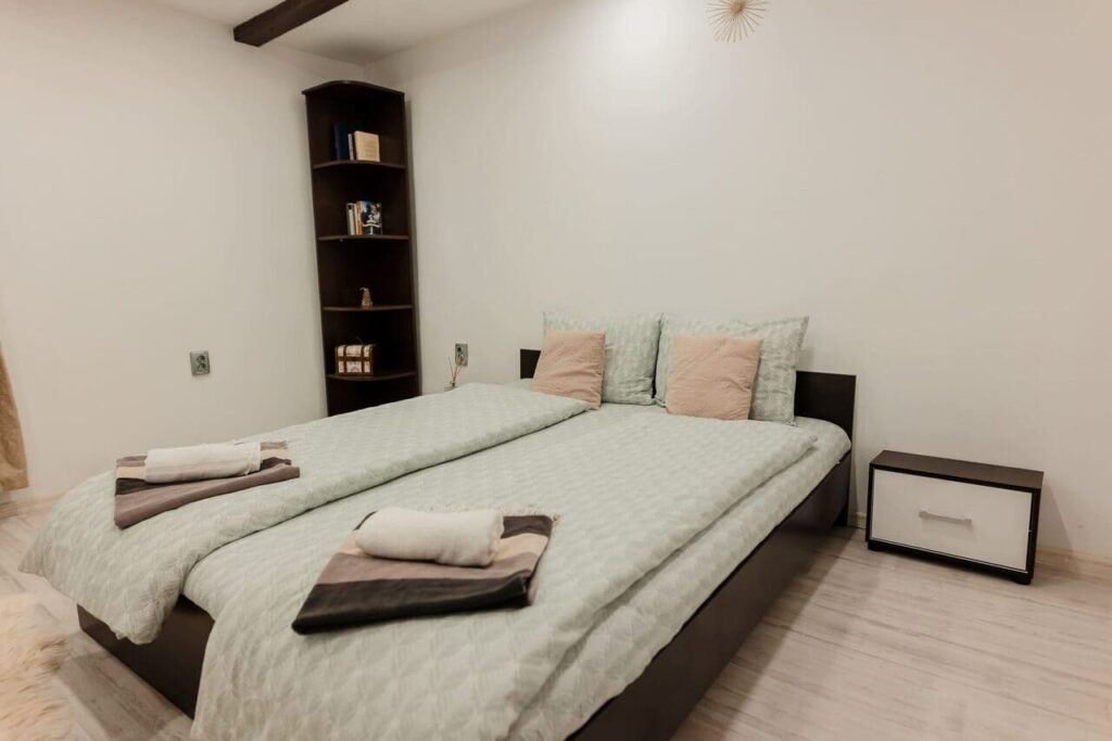 Imagine din Camera 1 de la parter, cu patul dublu aranjat pentru două persoane, o bibliotecă de colț din lemn închis la culoare și o noptieră modernă. Decorul este simplu, minimalist, cu perne roz și prosoape aranjate cu grijă pe pat.