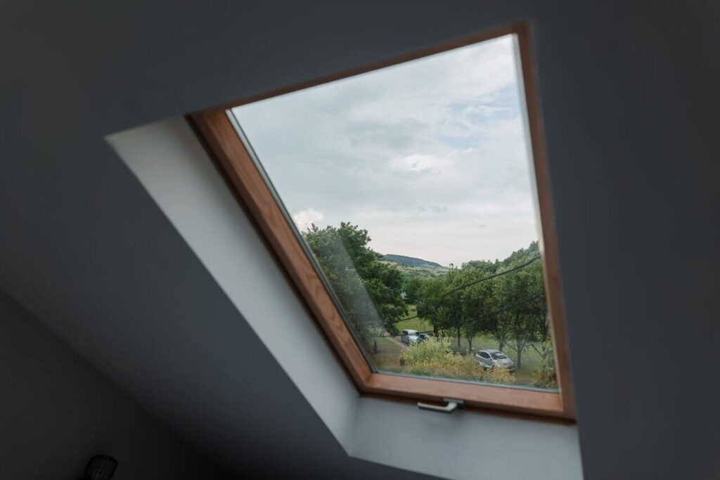 O imagine din interiorul Camerei 3 de la mansardă, cu un geam Velux deschis. Se observă o priveliște superbă a curții cabanei, cu copaci, spațiu verde și mașini parcate, sub un cer acoperit de nori. Fereastra aduce lumină naturală în cameră și permite oaspeților să admire peisajul.
