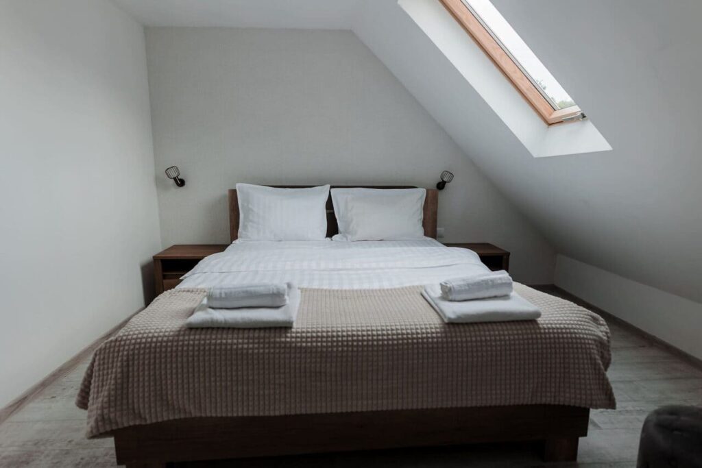 O imagine luminoasă a Camerei 3 de la mansarda Cabanei Ana. Se observă un pat matrimonial, cu lenjerie albă și cuvertură bej, două noptiere din lemn și un luminator Velux pe acoperiș. Design-ul este minimalist, cu pereți albi și o atmosferă aerisită.