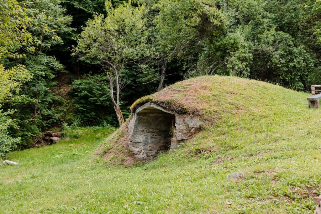 O imagine de aproape a intrării în crama veche, săpată în pământ și acoperită cu iarbă. Intrarea este din piatră, cu un aspect rustic și autentic, sugerând o construcție tradițională. Crama se află într-o zonă verde, cu vegetație.