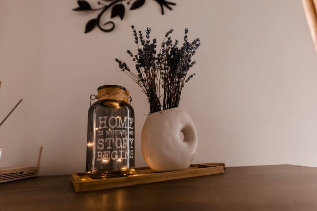 O fotografie de aproape a decorului de pe o comodă din livingul Cabanei Ana. Se observă un borcan de sticlă cu o lumină LED și mesajul "Home is where our story begins", alături de un vas ceramic alb cu flori uscate de lavandă, așezate pe o tavă de lemn.