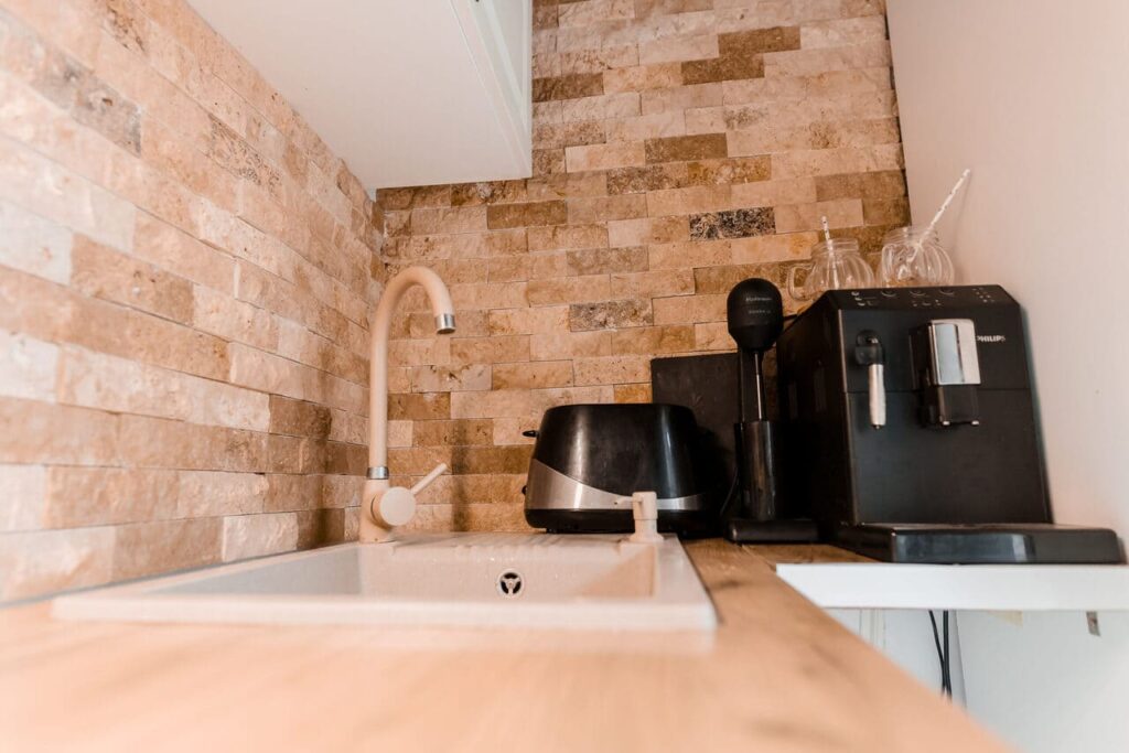 Un prim-plan al electrocasnicelor din bucătăria Cabanei Ana, care include un aparat de cafea Philips, un toaster, o chiuvetă modernă și un robinet elegant. Peretele din spatele blatului de lemn este decorat cu piatră naturală, adăugând un element rustic designului.