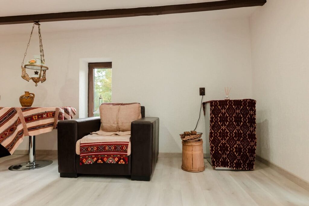 Un fotoliu maro, modern, decorat cu materiale textile cu motive tradiționale românești, amplasat într-un colț al Camerei 4 din anexa Cabanei Ana. Se observă și o măsuță rustică din lemn, precum și un mini-frigider acoperit cu o țesătură tradițională.