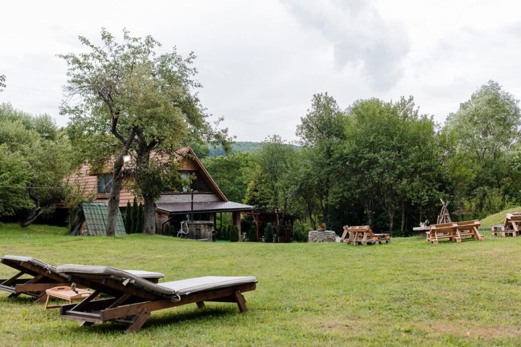 O imagine de ansamblu a Cabanei Ana și a grădinii, văzută dintr-un unghi mai înalt, de lângă foișor. Se observă șezlongurile din prim-plan, spațiul vast de iarbă, zona pentru foc de tabără și exteriorul cabanei principale. Grădina este înconjurată de copaci și vegetație.