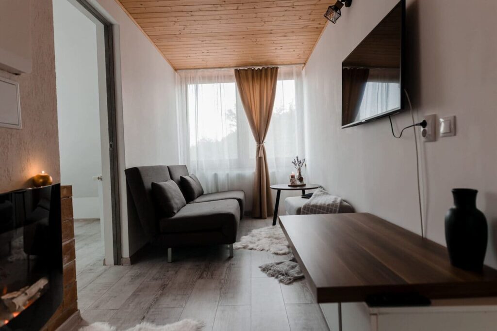 O imagine de ansamblu a zonei de relaxare de la etajul Cabanei Ana, cu o canapea extensibilă gri, o măsuță de cafea, un televizor montat pe perete și un șemineu electric în colțul camerei. Vederea este de la intrarea în cameră, spre fereastra mare cu perdele albe și bej.