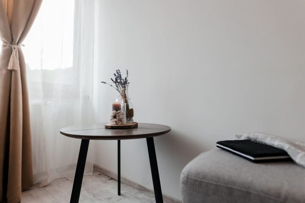 O măsuță de cafea minimalistă, cu picioare negre și blat rotund din lemn, amplasată în holul de la etajul Cabanei Ana. Pe măsuță se află un vas cu flori uscate de lavandă și o lumânare, creând o atmosferă relaxantă. În plan îndepărtat se observă o perdea elegantă și o canapea gri.