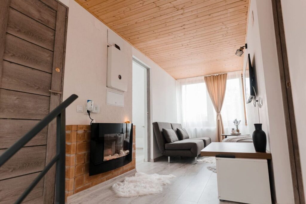 O imagine de ansamblu a holului de la etajul Cabanei Ana, cu un șemineu electric modern în prim-plan. Se observă o canapea gri, un televizor, o ușă de lemn și o scară cu balustradă metalică. Tavanul este lambrisat cu lemn deschis la culoare.