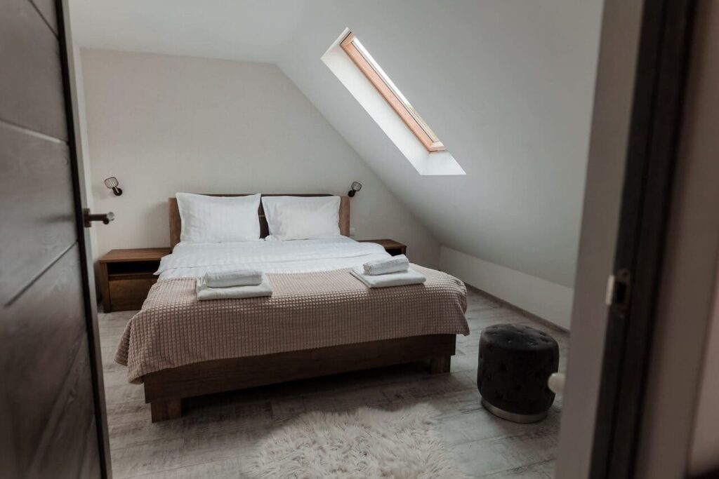 O vedere din holul de la etaj spre interiorul Camerei 3. Se observă un pat dublu, un luminator pe acoperiș și o noptieră modernă. Ușa de la intrare se deschide spre un spațiu curat, cu design minimalist, care invită la relaxare.