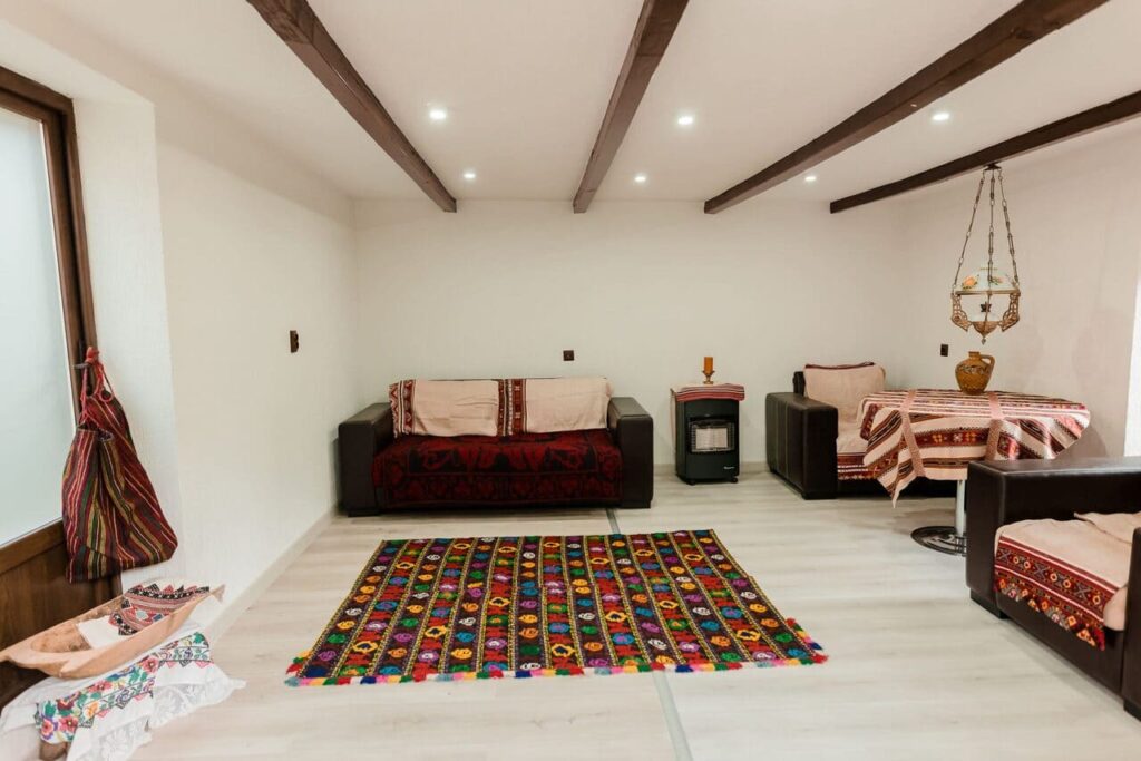 Vedere de la ușă în interiorul Camerei 4 din anexa Cabanei Ana. Se observă mobilier modern, canapele confortabile, o masă de servit masa și obiecte decorative tradiționale, cum ar fi un covor multicolor și o lampă rustică. Tavanul este decorat cu grinzi din lemn masiv.