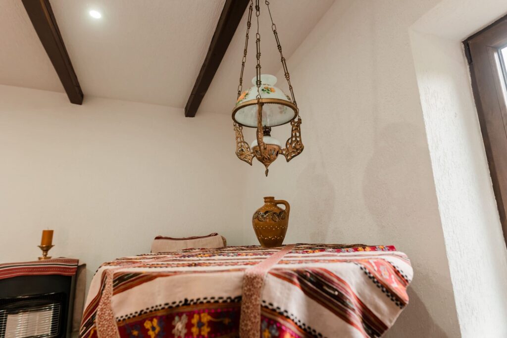 Masa din Camera 4, acoperită cu o față de masă cu motive tradiționale românești, decorată cu un vas din lut. Deasupra mesei atârnă o lampă suspendată, cu un design vintage și detalii ornate, care completează atmosfera autentică a încăperii.
