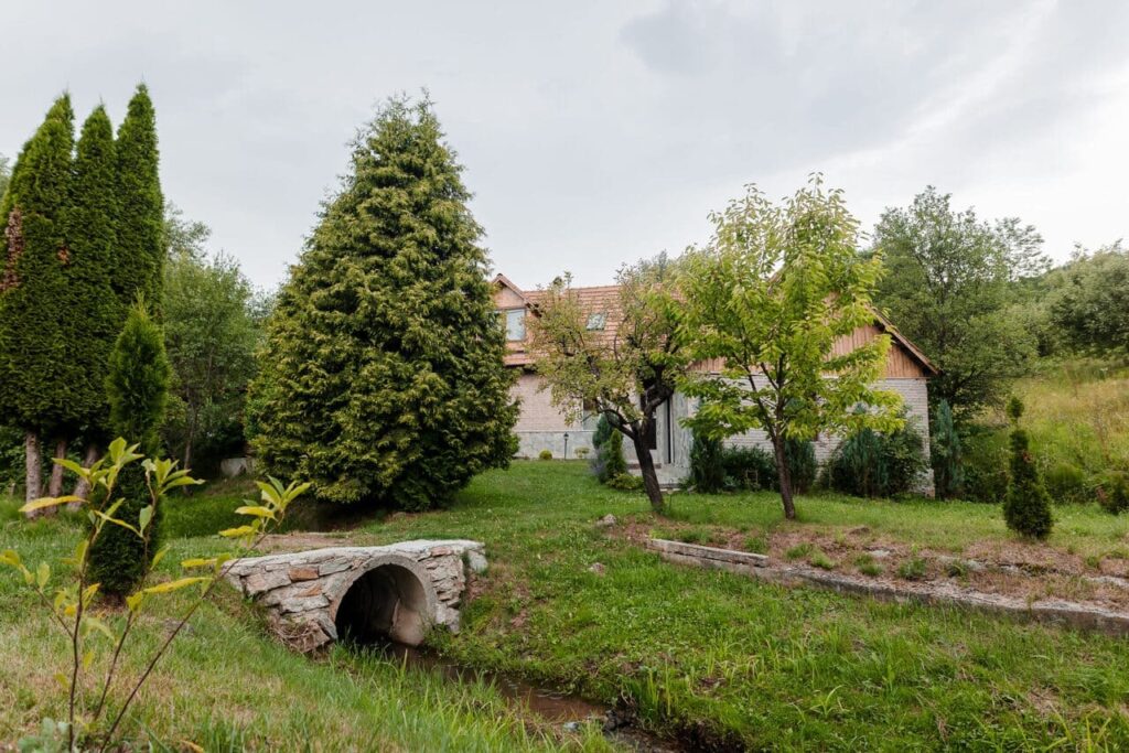 O imagine cu un pod de piatră rustică peste un pârâu mic, care asigură accesul spre anexa Cabanei Ana. Se observă anexa cu pereți din piatră, copaci verzi și un peisaj natural, care subliniază armonia și frumusețea proprietății.