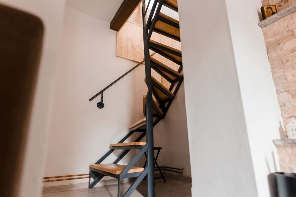 Scări interioare spiralate, cu trepte din lemn și balustradă metalică, care fac legătura între parter și etajul Cabanei Ana. Se observă un design modern, care economisește spațiu, alături de pereții albi și un perete decorativ cu piatră, pe fundal.
