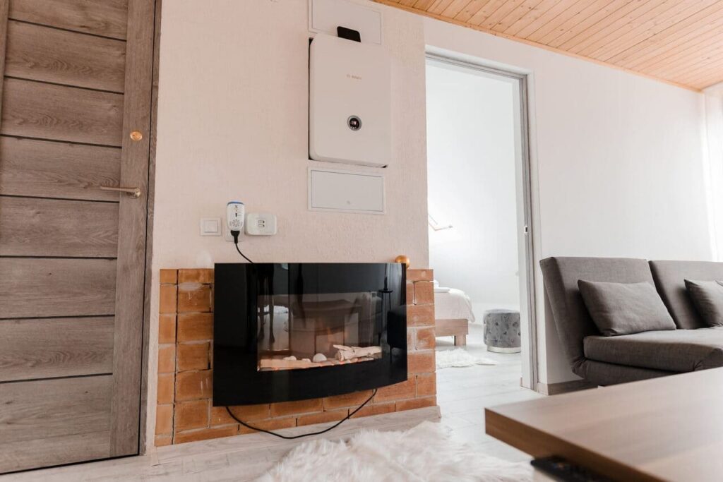 O imagine de aproape a șemineului electric din holul de la etajul Cabanei Ana. Șemineul este încastrat într-un perete cu placaj de cărămidă, iar deasupra se află centrala termică și un termostat. Se observă o canapea gri pe fundal și o ușă de lemn închis la culoare.