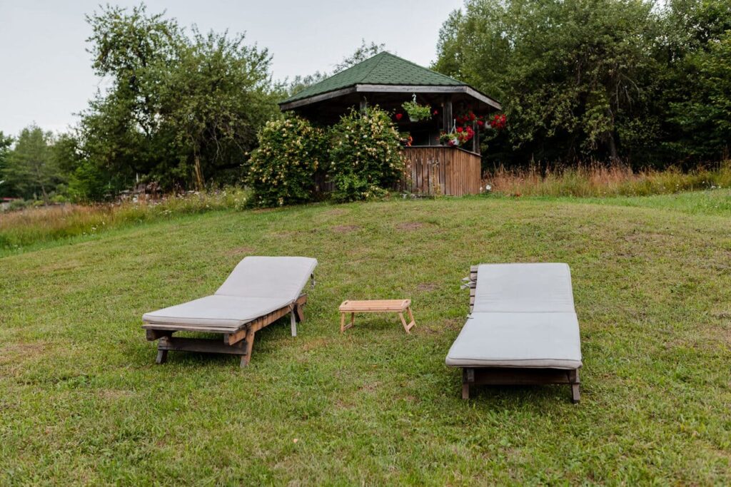 O imagine din grădina Cabanei Ana, cu două șezlonguri din lemn, așezate pe o zonă cu iarbă. În fundal se observă un foișor din lemn cu acoperiș verde și mese, ideal pentru relaxare și socializare.