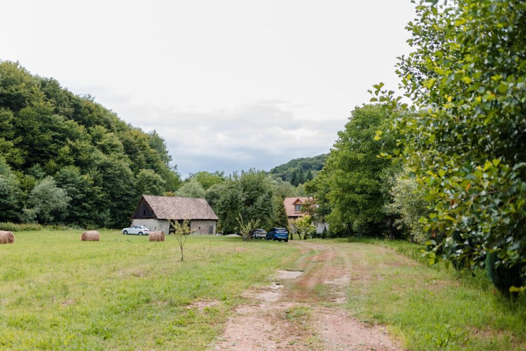 O imagine de la intrarea pe proprietatea Cabanei Ana, cu un drum de pământ care se deschide spre un spațiu verde imens. În stânga se află o clădire rustică, o sură veche cu acoperiș din țiglă, iar în dreapta se vede o parte din cabană. Peisajul este înconjurat de copaci și dealuri.