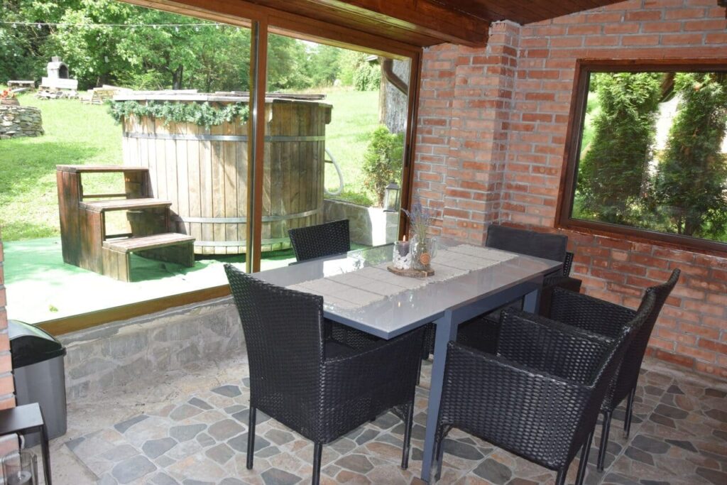 O imagine din interiorul terasei acoperite de la Cabana Ana. Se observă o masă de dining elegantă, cu scaune negre, și o priveliște directă spre ciubărul din lemn situat în grădină. Pereții din cărămidă și geamurile mari creează o atmosferă luminoasă și primitoare.