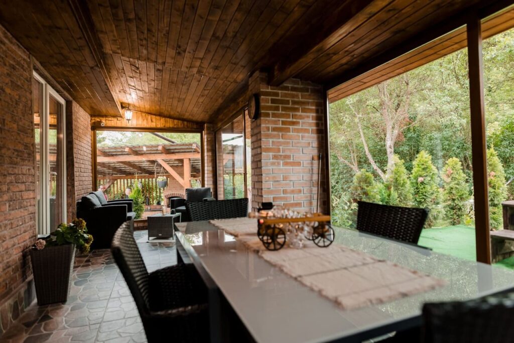 Imagine din interiorul terasei acoperite de la Cabana Ana, cu o masă mare, scaune elegante, și o canapea confortabilă. Se observă tavanul din lemn și pereții din cărămidă, cu o priveliște directă spre grădina verde și luxuriantă a proprietății.