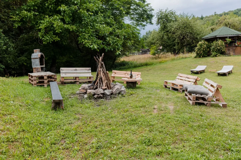 Vedere de ansamblu a zonei pentru foc de tabără de la Cabana Ana, Feleacu. Imaginea prezintă un spațiu circular, cu bănci din lemn din paleți și scaune, așezate în jurul unui loc special amenajat pentru foc, în mijlocul unei grădini pline de verdeață și copaci.