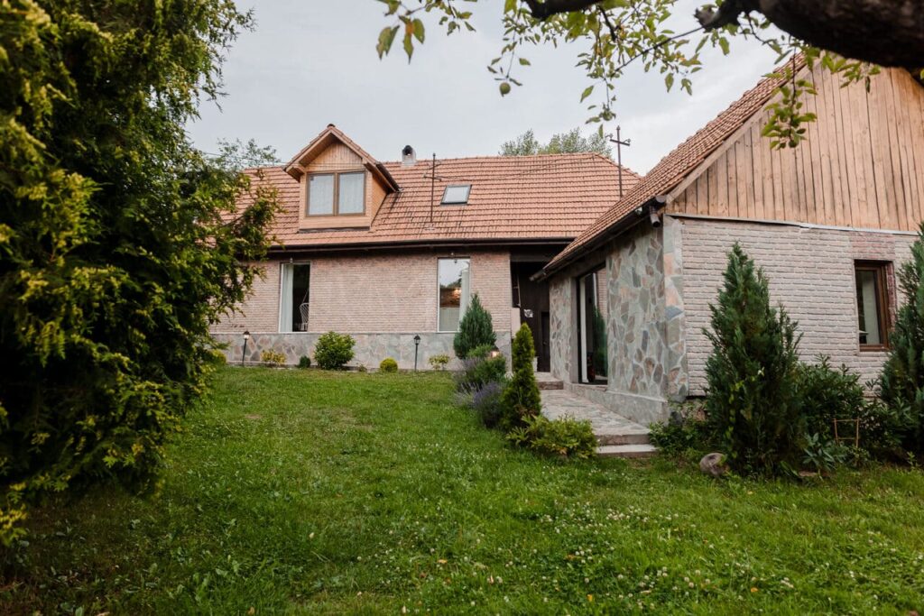 O imagine de ansamblu, din lateral, a Cabanei Ana și a noii anexe. Se observă arhitectura clădirilor cu pereți din cărămidă și piatră, ferestre mari și o zonă verde, cu vegetație decorativă, în fața clădirilor.